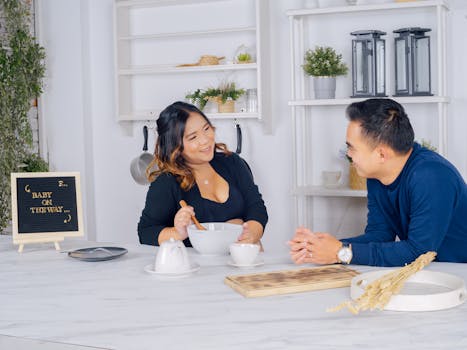 Asian couple in a kitchen setting with a pregnancy announcement sign 'Baby on the Way', smiling at each other.