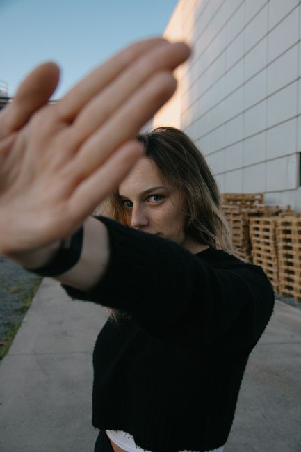 Young female in casual clothes closing camera with hand while standing on pavement near building and looking at camera