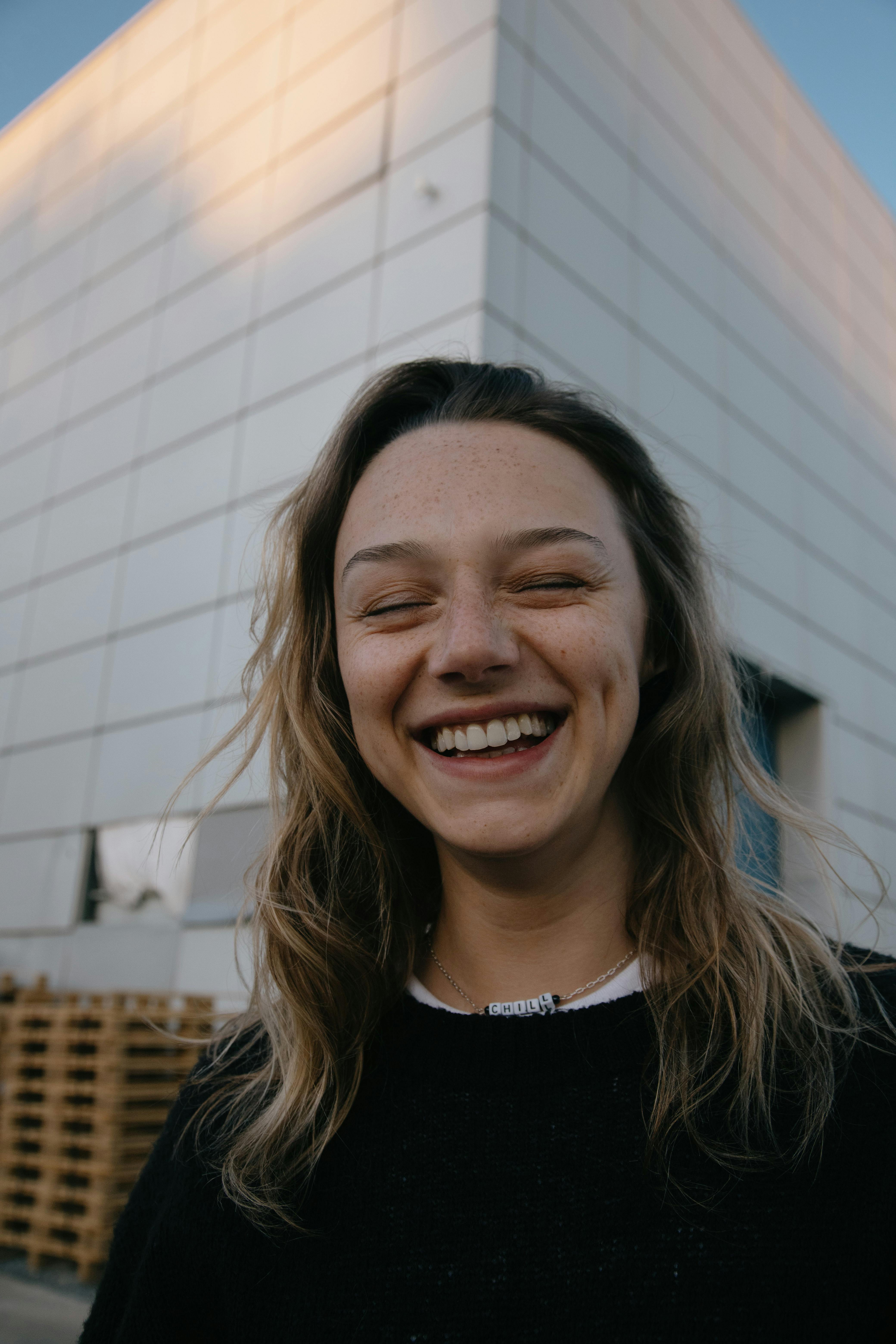 Young happy female with wavy hair and casual outfit laughing with closed eyes while standing near urban building