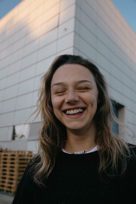 Young happy female with wavy hair and casual outfit laughing with closed eyes while standing near urban building