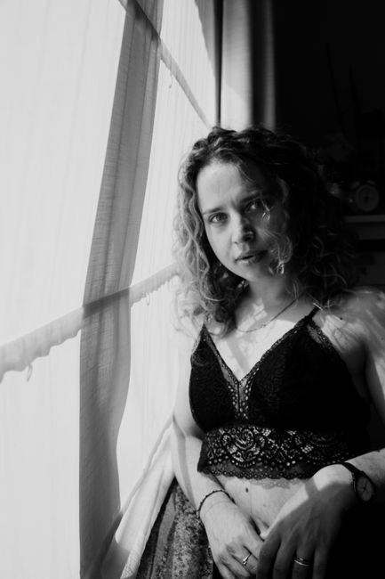 Black and white of young attractive female with curly hair in lace lingerie standing near window and looking at camera