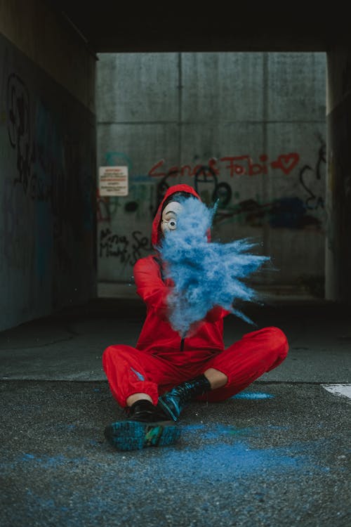 Full body of person in unusual outfit and scary mask sitting on ground and throwing blue powder