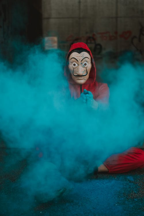 Person in scary mask and hood sitting on ground covered with color powder
