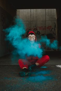 小巷里，一个身穿红衣、戴着面具的人坐在那儿，四周弥漫着鲜艳的蓝烟