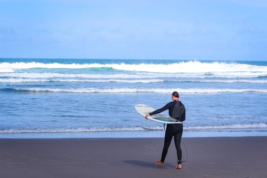 A surfer in a wetsuit carrying a surfboard along the sandy Piha Beach, New Zealand.