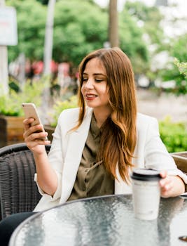 Smiling woman in a blazer uses smartphone while enjoying coffee outdoors in Guadalajara.