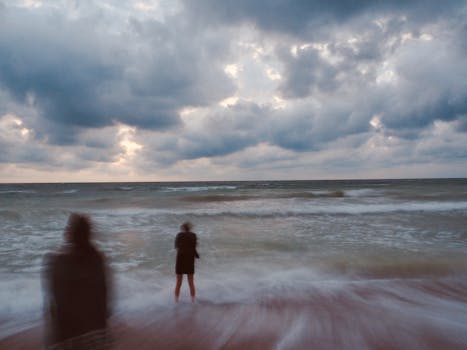 Motion-blurred silhouettes on a beach with dramatic clouds at twilight, capturing a serene yet moody atmosphere.