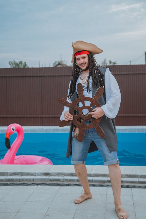 Full body of joyful young hotel animator in costume of pirate with artificial steering wheel having fun near swimming pool on cloudy day