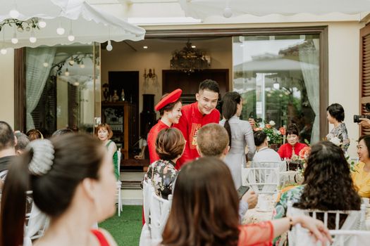 Smiling ethnic groom and bride in traditional red clothes speaking with unrecognizable guests during banquet