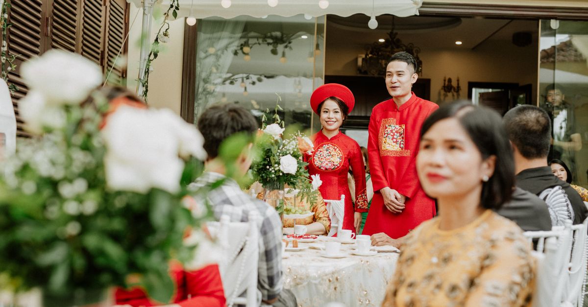 Smiling ethnic husband and wife in traditional red apparel near table with decor and anonymous guests during wedding banquet