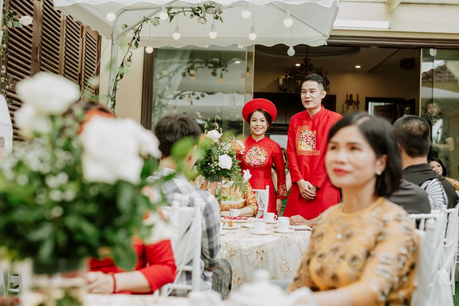 Smiling ethnic husband and wife in traditional red apparel near table with decor and anonymous guests during wedding banquet