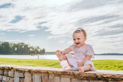 Free Full length happy smiling girl wearing white summer dress sitting on stone fence on waterfront on sunny day Stock Photo