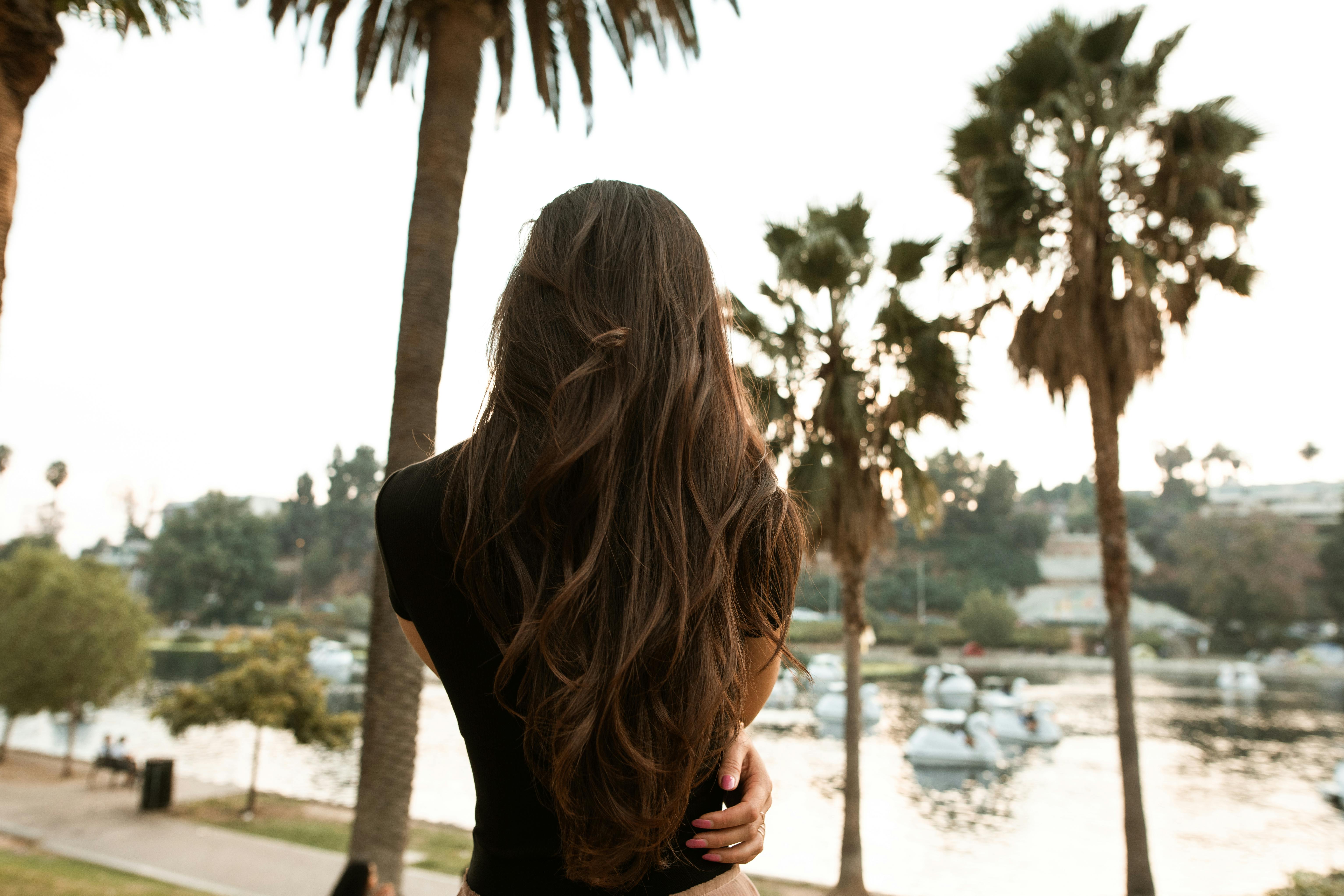 一名长发女子站在湖边，背影周围是风景优美的棕榈树
