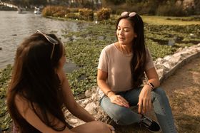 Two women enjoying a casual conversation by a tranquil lakeside in warm daylight.
