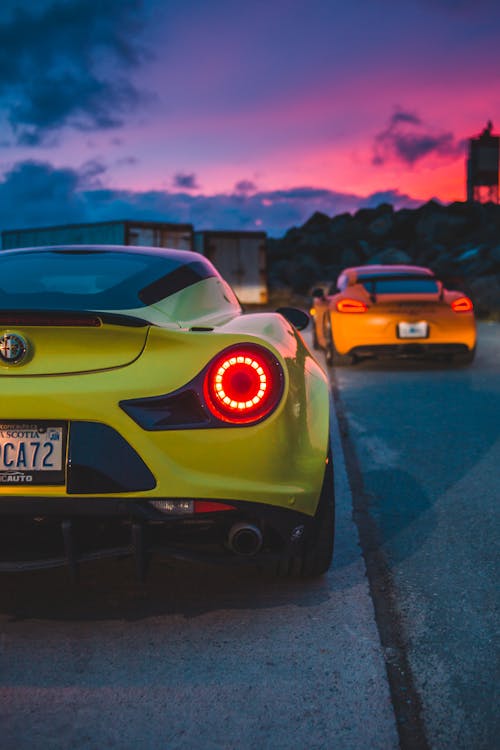 Luxury contemporary sports cars with red tail lights driving on asphalt roadway under colorful sky at twilight