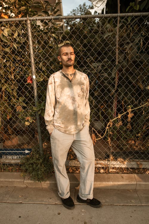 無料 金網フェンスの横に立っている灰色のドレスシャツと白いズボンの男 写真素材