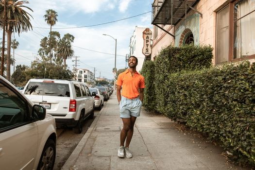 Stylish man in orange shirt and shorts walking on a sunny city street, smiling.