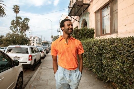 Happy man in bright orange shirt walks on a sunny day along a city street lined with hedges.