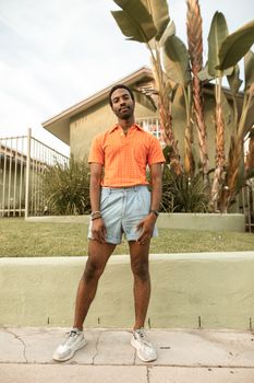 A fashionable man stands confidently outdoors in denim shorts and an orange polo shirt.