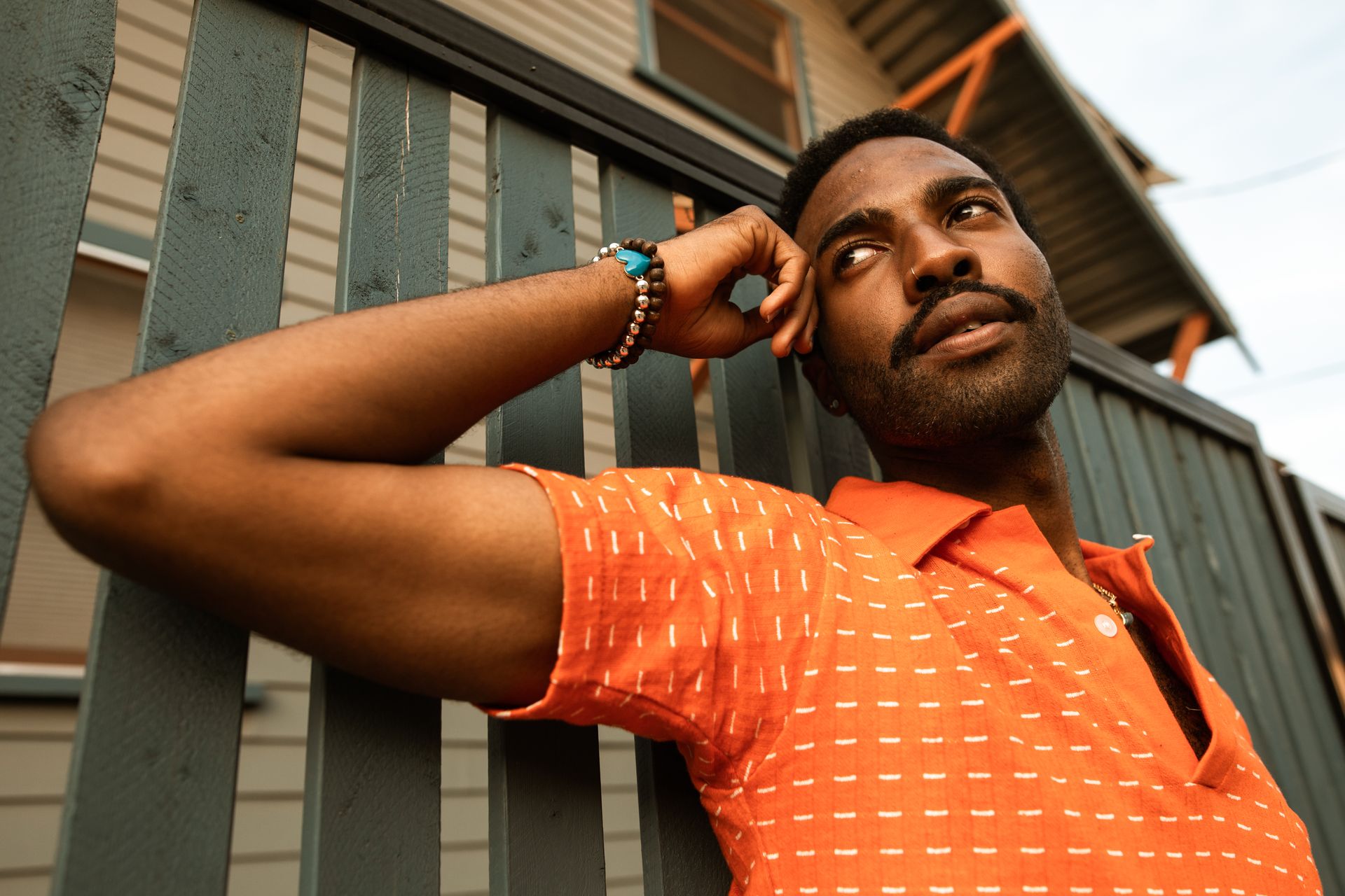 Close-up of a man in an orange polo shirt leaning against a wooden fence on a sunny day.