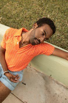 Portrait of a handsome man in an orange polo sitting outdoors on pavement with grass background.