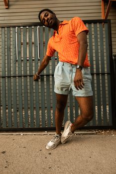 Stylish young man posing in an orange polo shirt and denim shorts against a modern fence.