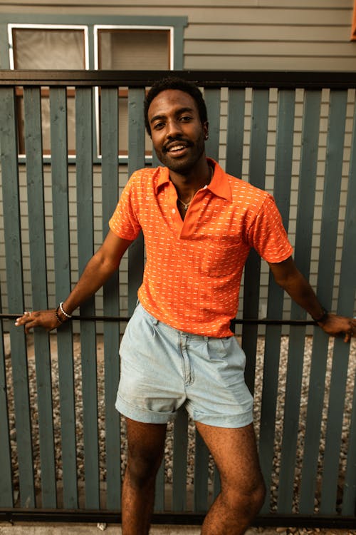 Man in Orange Polo Shirt Posing on Sidewalk by Fence · Free Stock Photo