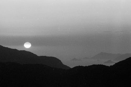 Black and white of minimal landscape with circle of white sun setting behind black mountain and ocean