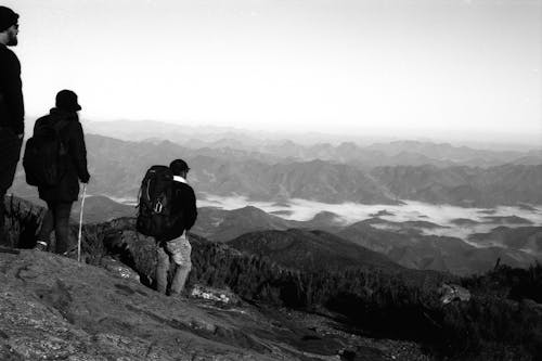 Black and white tourists with trekking poles standing on high slope of mountain admiring majestic ridge