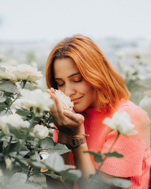 Attractive female with closed eyes smelling white flower while standing near blooming bush in garden with plants on blurred background