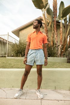 Fashionable man in orange shirt and shorts standing outdoors, looking away.