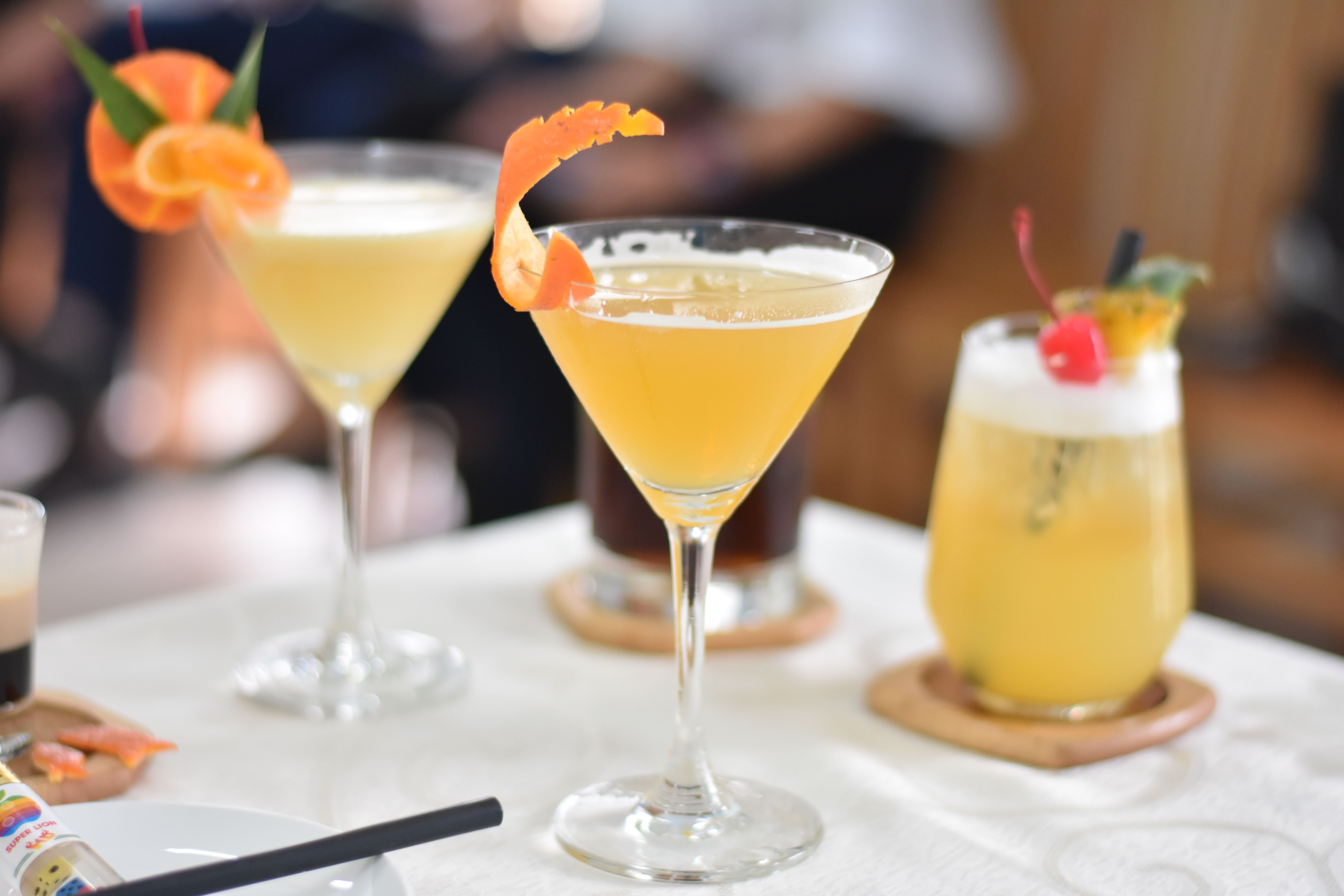 Close-up of diverse cocktails with orange and cherry garnishes on a white table.