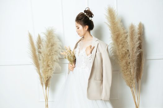 Bride in a white dress with a bouquet, standing indoors alongside pampas grass.