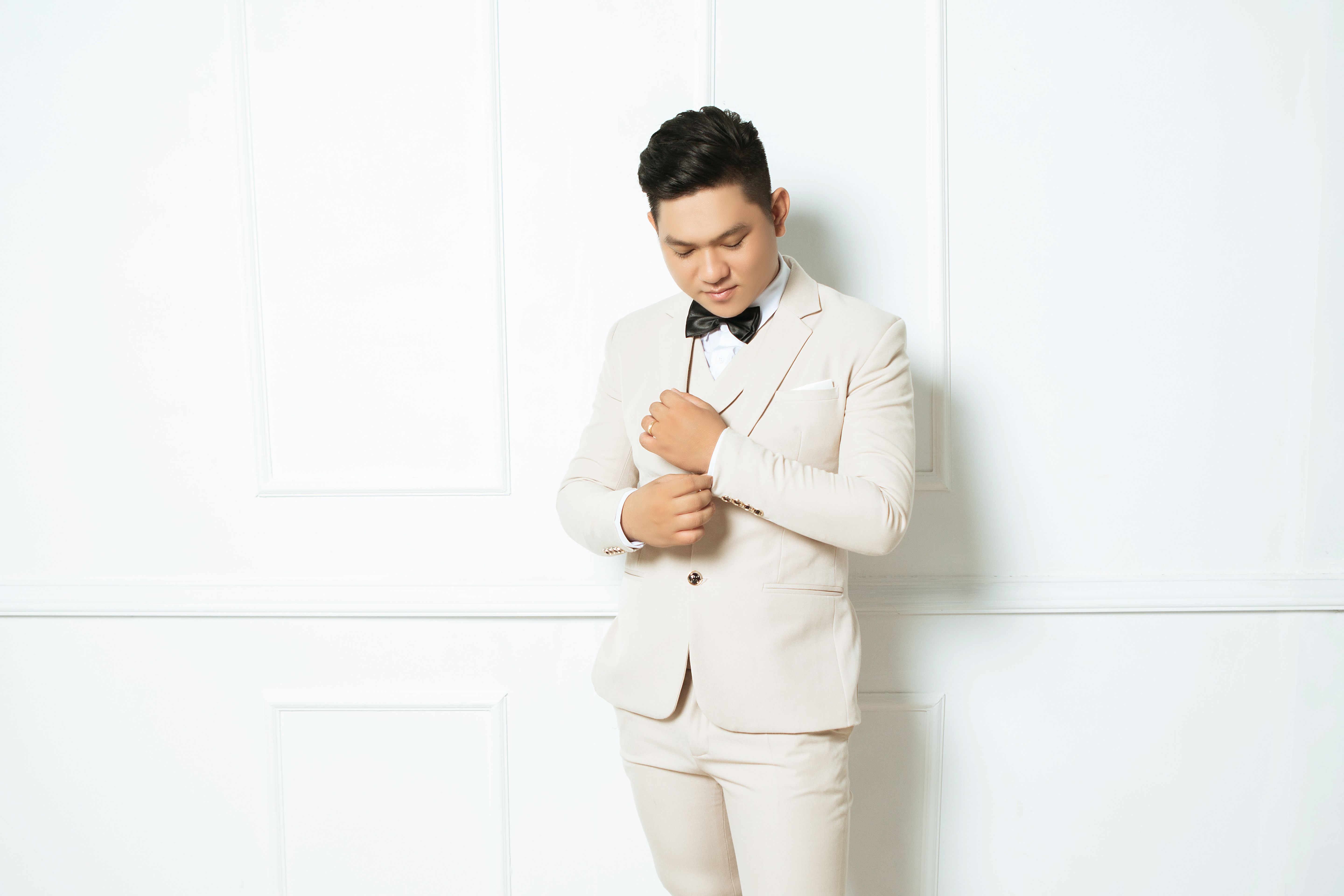 A stylish groom in a light suit adjusting his bow tie against a white indoor background.