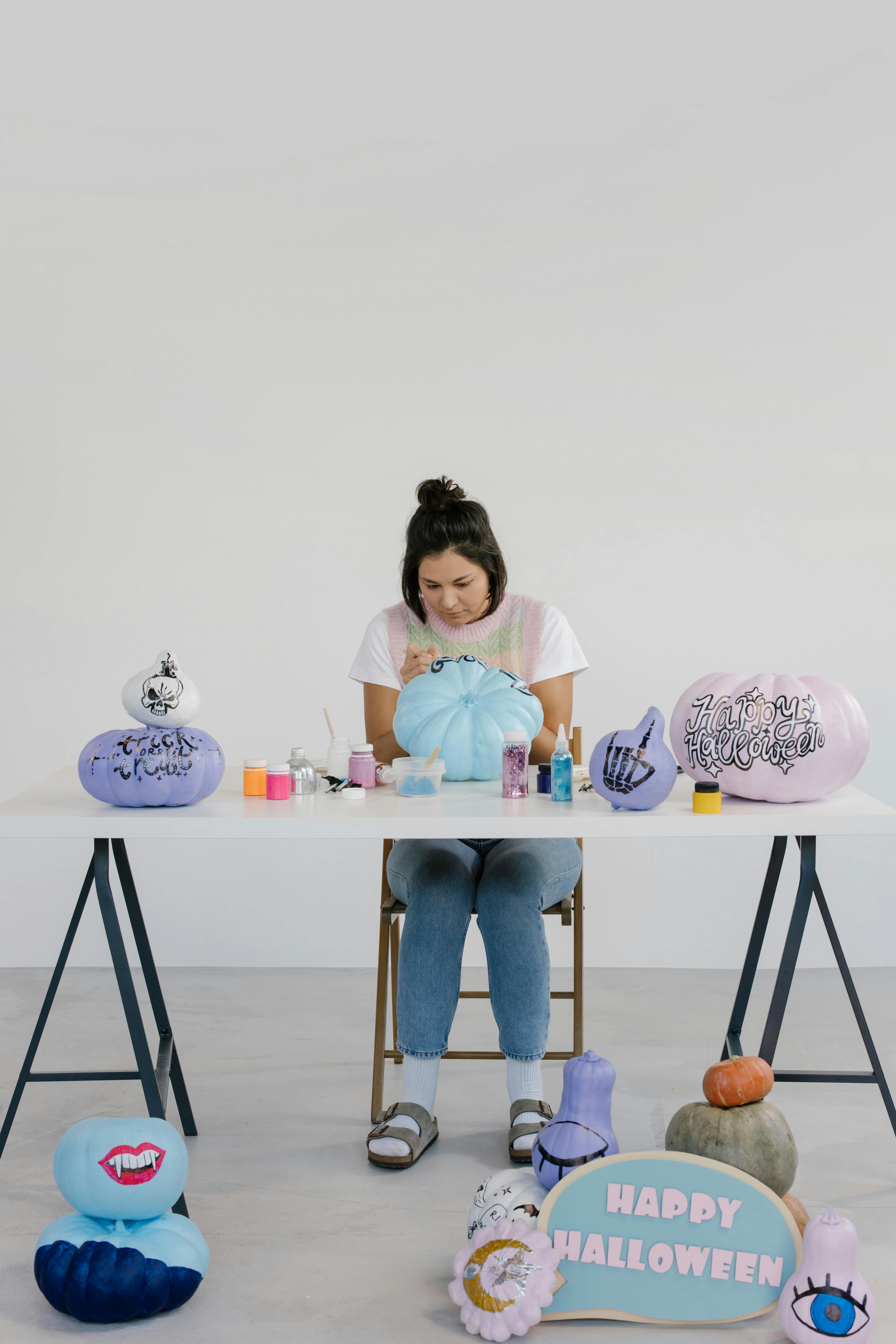 woman in white shirt sitting on white wooden table while painting a pumpkin