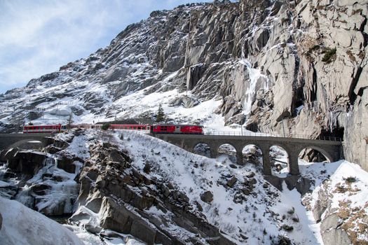 A vibrant red train traverses a scenic snowy viaduct in the Swiss Alps.