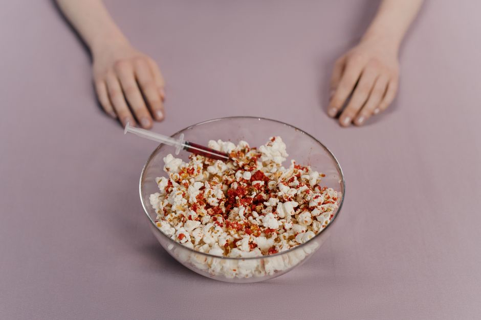Close-up of popcorn in a glass bowl with a syringe on a pink surface, concept art.