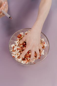 A hand mixes popcorn with a unique red topping using a syringe against a purple background.