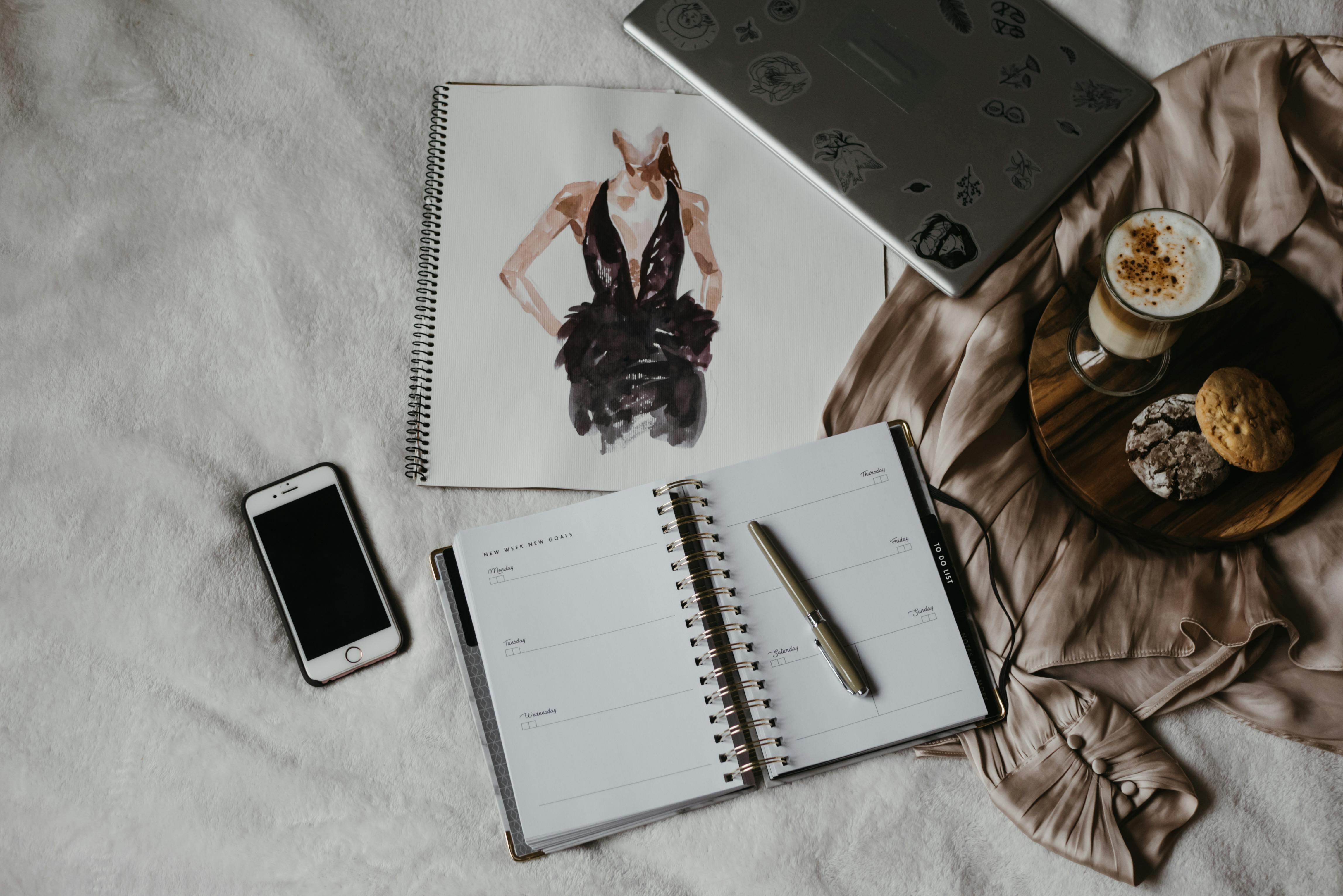 Flat lay of a creative workspace featuring a planner, coffee, smartphone, and fashion sketch.