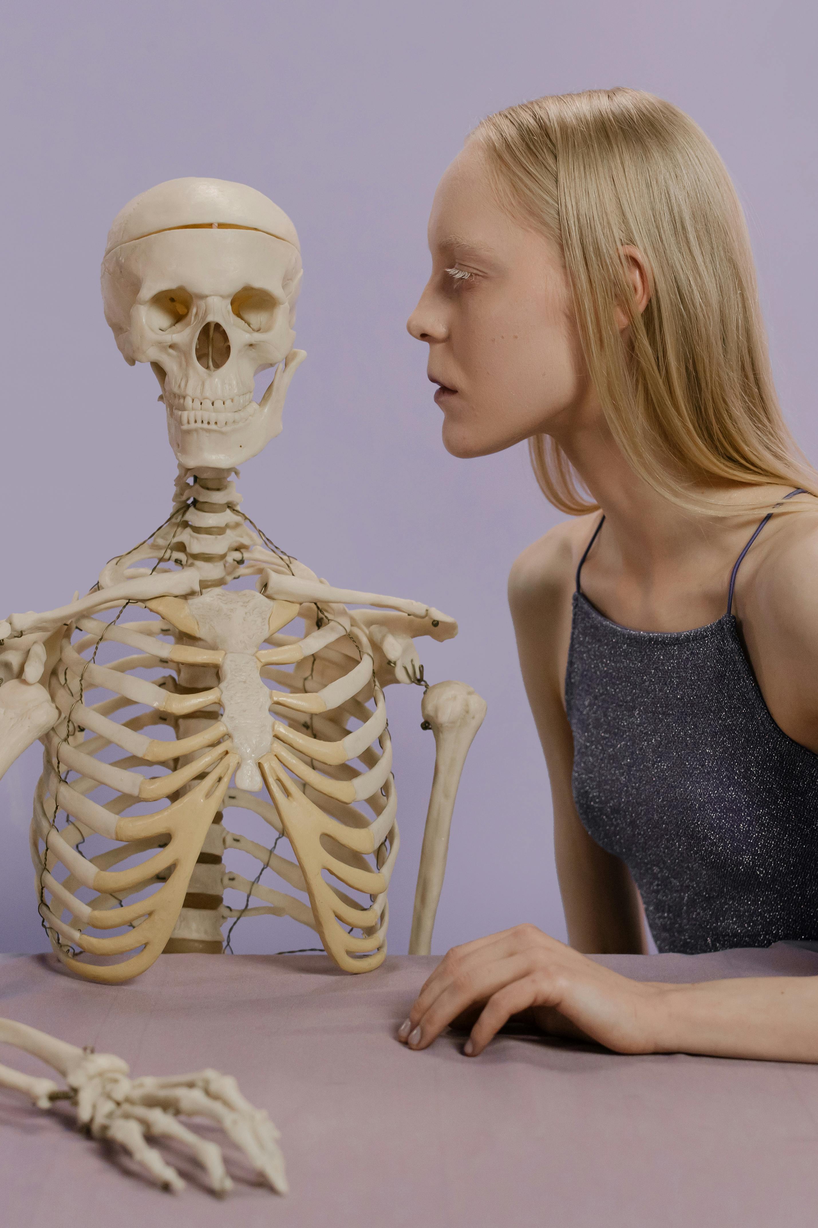 A woman poses creatively with a skeleton in a studio setting, highlighting anatomy and artistic expression.