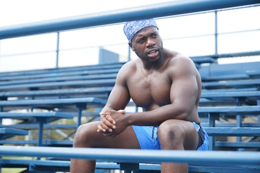 Shirtless muscular man sitting on outdoor stadium bleachers, looking focused.