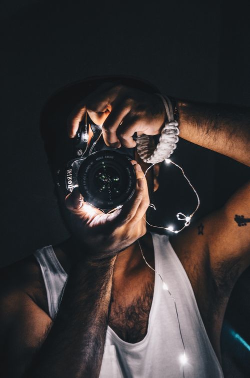 Free Artistic portrait of a male photographer using a camera, wrapped in string lights, in a dark setting. Stock Photo