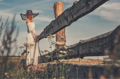 Free Woman in White Off Shoulder Dress and Black Sun Hat Standing Behind a Wooden Fence Stock Photo