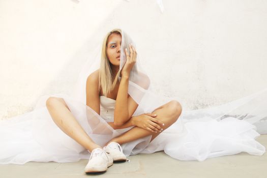 Blond Woman in Wedding Dress and Snickers Sitting on Concrete Pavement