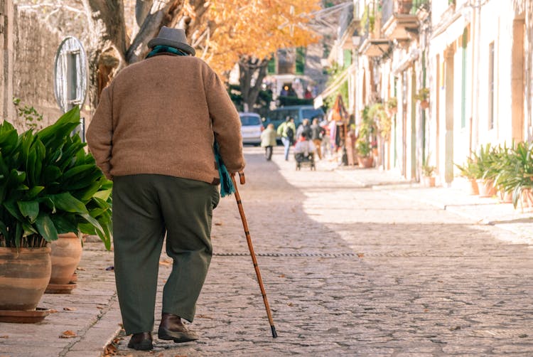 Elderly Man In Brown Sweater Walking On Stone Pavement With A Cane