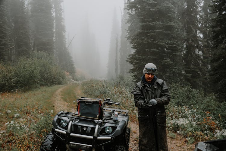 Male Traveler In Raincoat On Path Near ATV Between Woods