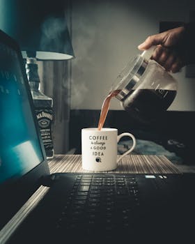 A hand pours hot coffee into a ceramic mug near a laptop, creating a warm, cozy atmosphere.