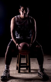 A young male basketball player sitting on a stool in a dimly lit environment.