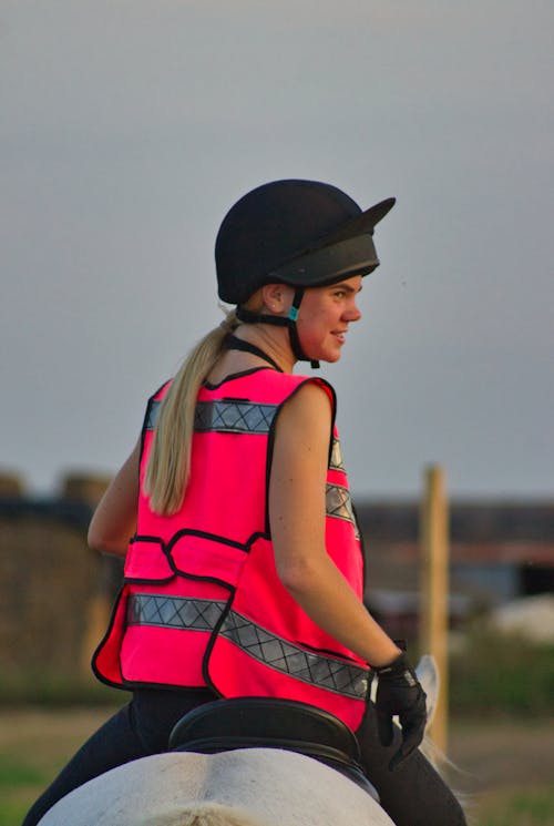 Back view of positive young female equestrian in bright vest and helmet sitting on gray horse and looking away on sunny day in countryside