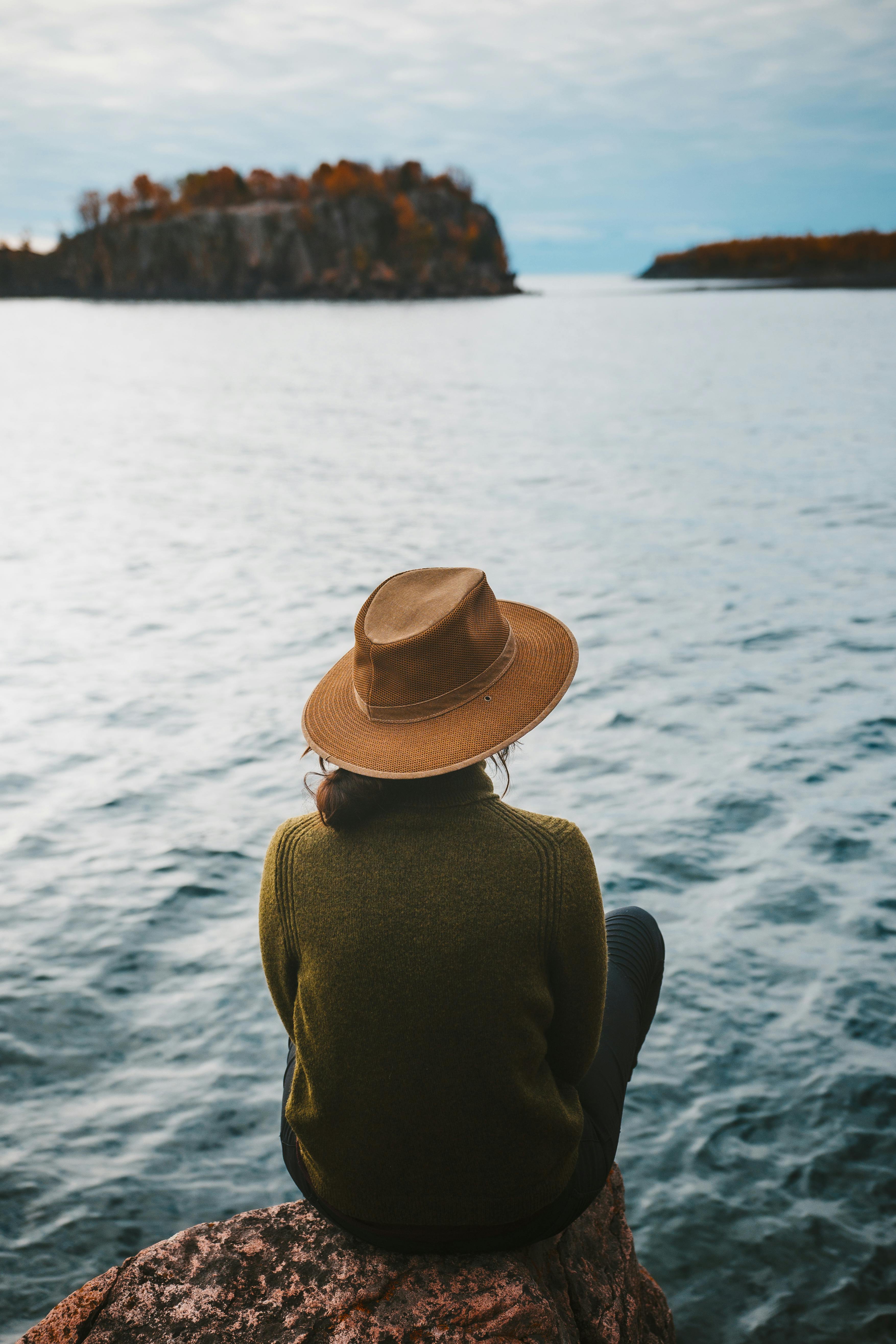 back view of a person in brown cowboy hat and green sweatshirt sitting on a cliff overlooking the seascape scenery